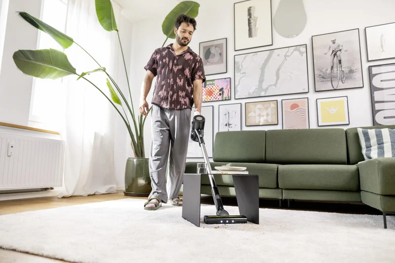 Person mit Kärcher Staubsauger in modernem Wohnzimmer, grüne Couch, Pflanzen, Wandbilder, heller Teppich.
