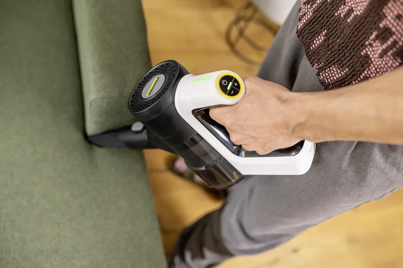 Person reinigt grünes Sofa mit einem Kärcher Handstaubsauger in einem Raum mit Holzboden.