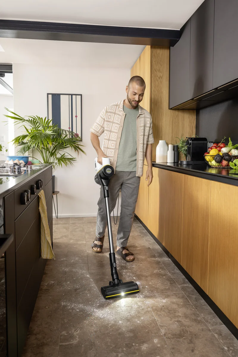 Mann reinigt Küchenboden mit einem Kärcher Dampfreiniger. Moderne Küche mit Holzschränken und Obstschale auf der Arbeitsplatte.