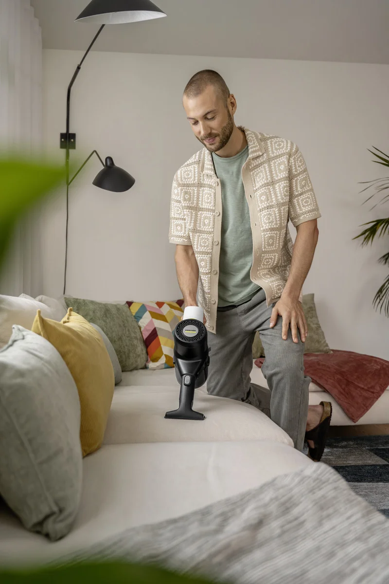 Person reinigt ein Sofa mit einem Kärcher Handstaubsauger in einem Wohnzimmer. Kissen und eine Stehlampe sind im Hintergrund sichtbar.