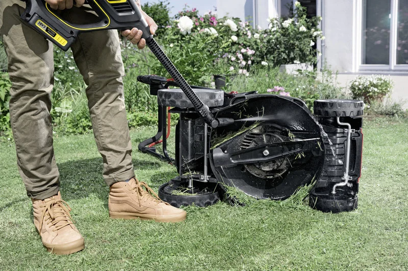Person reinigt Rasenmäher mit Kärcher Hochdruckreiniger im Garten. Der Mäher ist umgedreht, Grasreste sind sichtbar.