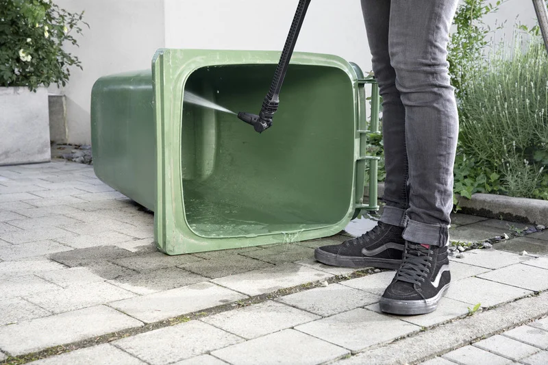 Person reinigt grünen Müllcontainer mit Kärcher Hochdruckreiniger auf gepflastertem Boden im Freien.