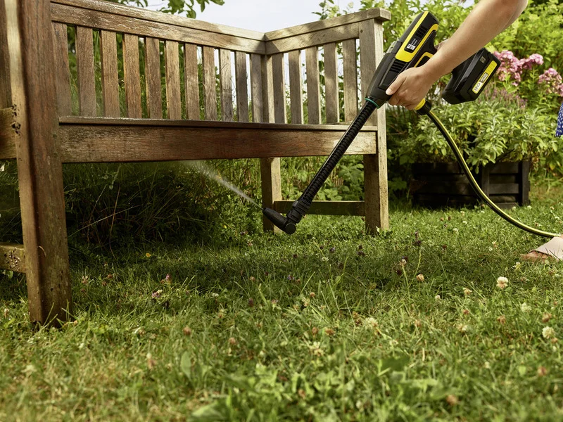 Person reinigt Holzbank im Garten mit Kärcher Hochdruckreiniger. Blumen und Gras im Hintergrund.