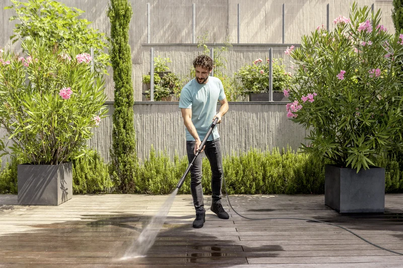 Person reinigt Terrasse mit Kärcher Hochdruckreiniger, umgeben von blühenden Oleanderpflanzen und einer modernen Gartenmauer.