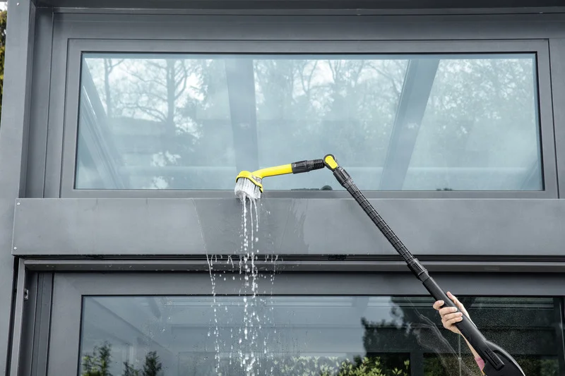 Person reinigt Fenster mit Kärcher Waschbürste, Wasser fließt herunter. Außenansicht eines modernen Gebäudes.