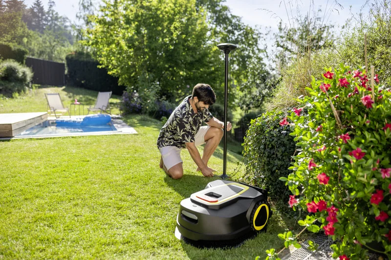 Mann im Garten neben einem Mähroboter, umgeben von blühenden Sträuchern, im Hintergrund ein Pool mit Liegestühlen.