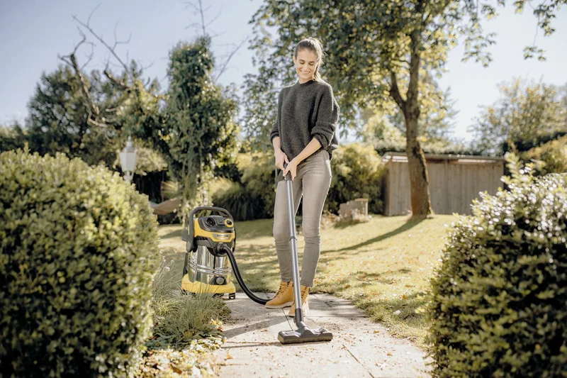 Person mit Kärcher Nass-/Trockensauger im Garten, umgeben von Büschen und Bäumen, sonniges Wetter.