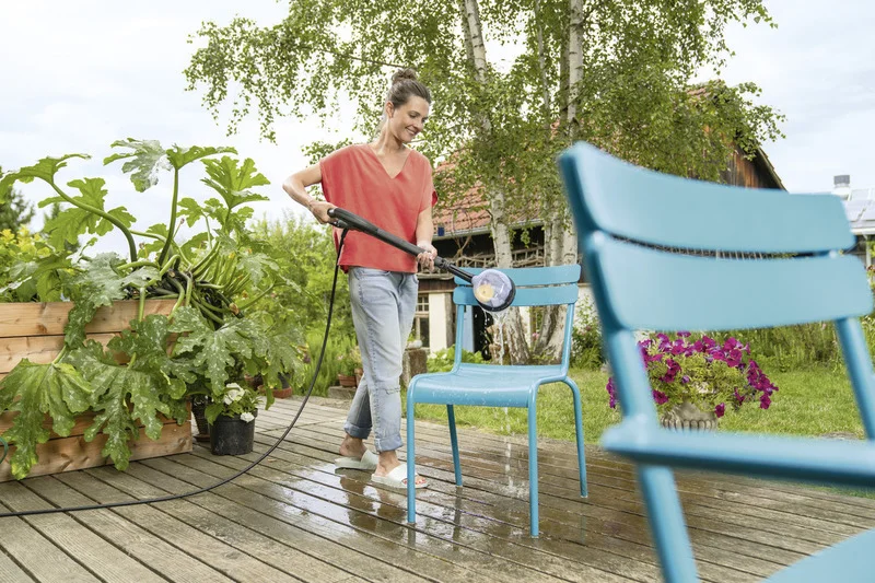 Person reinigt blauen Gartenstuhl mit Kärcher Hochdruckreiniger auf Holzterrasse, umgeben von Pflanzen und Bäumen.