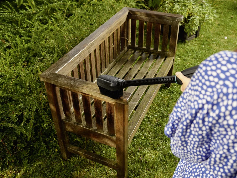 Holzbank im Garten wird mit Kärcher Reinigungsbürste gesäubert. Person in blau gepunktetem Kleid hält die Bürste.