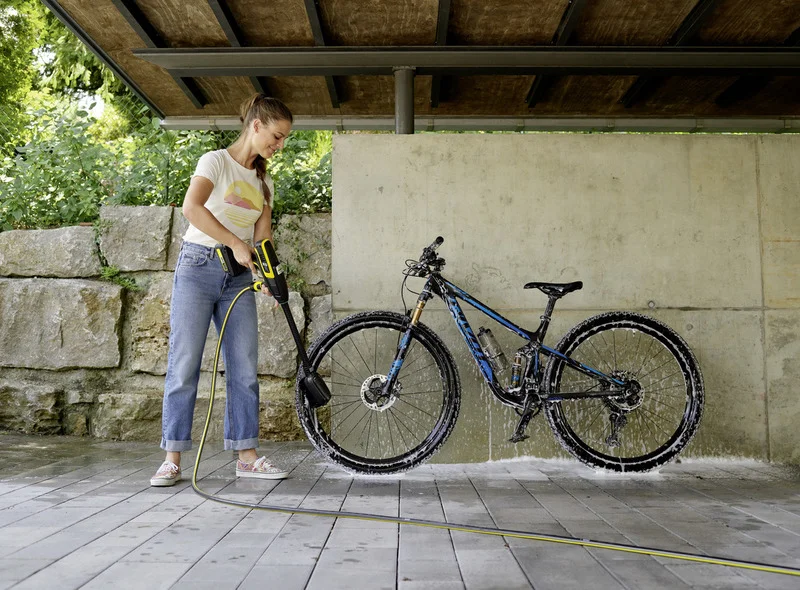 Person reinigt Fahrrad mit Kärcher Hochdruckreiniger unter überdachtem Bereich, umgeben von Steinmauer und grüner Vegetation.