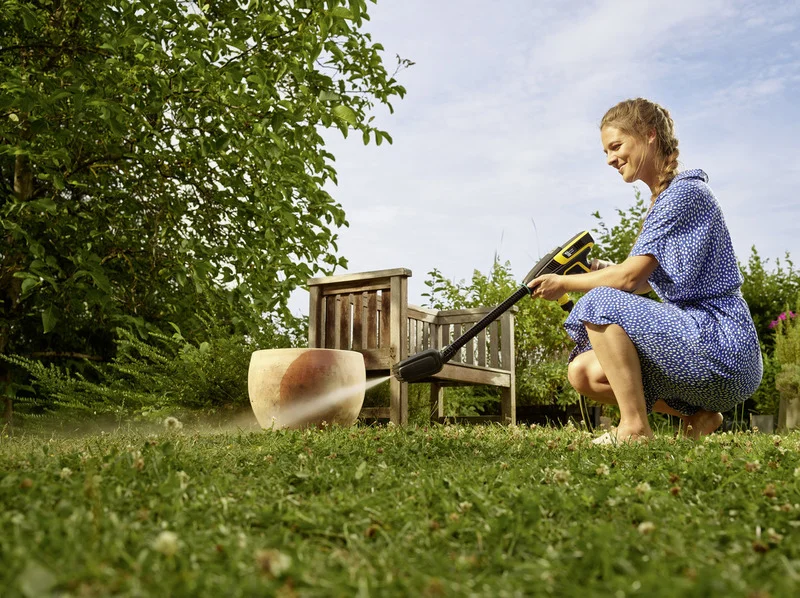 Frau reinigt einen großen Blumentopf im Garten mit einem Kärcher Hochdruckreiniger. Im Hintergrund sind Bäume und eine Holzbank.