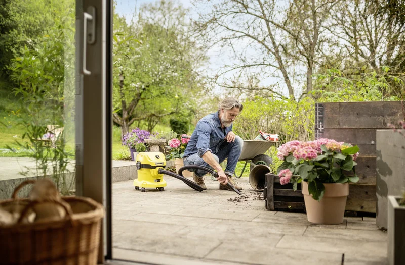 Mann reinigt Terrasse mit Kärcher Nass-Trockensauger, umgeben von Pflanzen und einer Schubkarre, im Garten bei sonnigem Wetter.