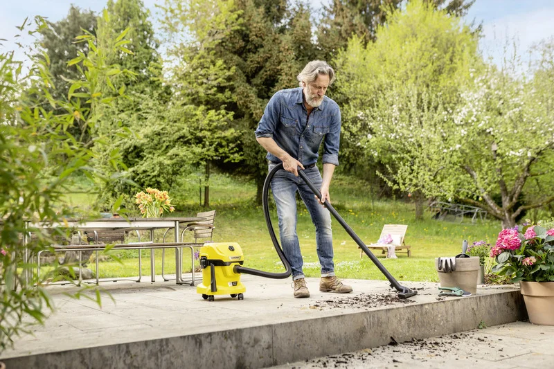 Mann reinigt Terrasse mit Kärcher Nass-/Trockensauger. Im Hintergrund Garten mit Tisch, Stühlen und blühenden Pflanzen.
