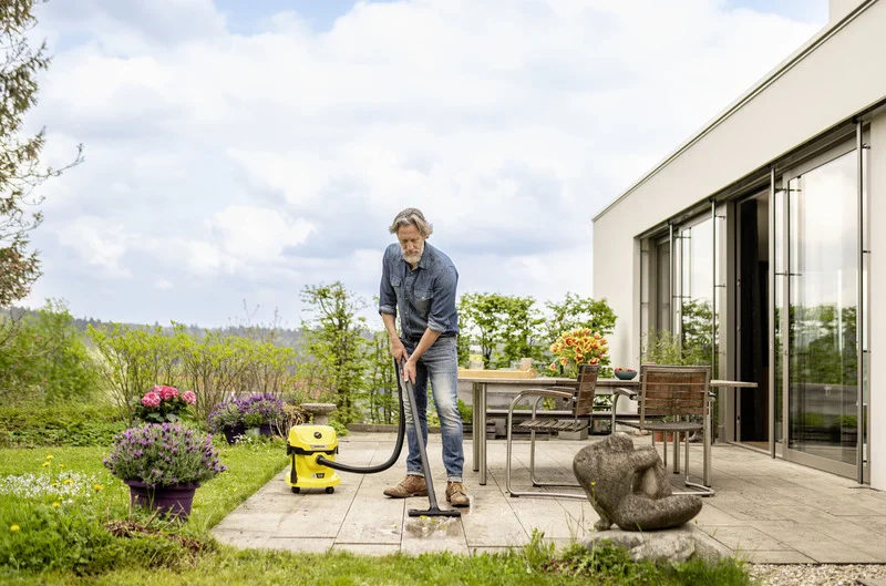 Person reinigt Terrasse mit Kärcher Nass-Trockensauger. Gartenmöbel und Blumen im Hintergrund, modernes Haus mit großen Fenstern.