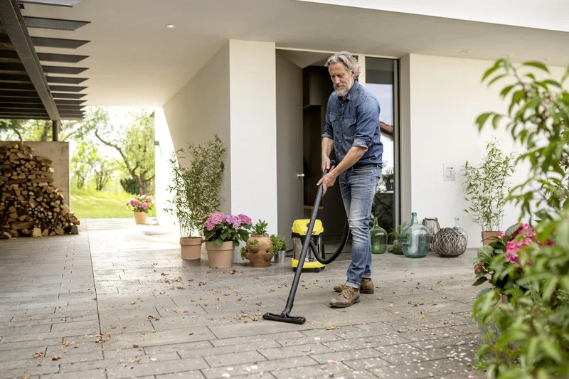 Mann reinigt Terrasse mit Kärcher Nass-Trockensauger, umgeben von Pflanzen und Holzstapel. Outdoor-Szene bei Tageslicht.