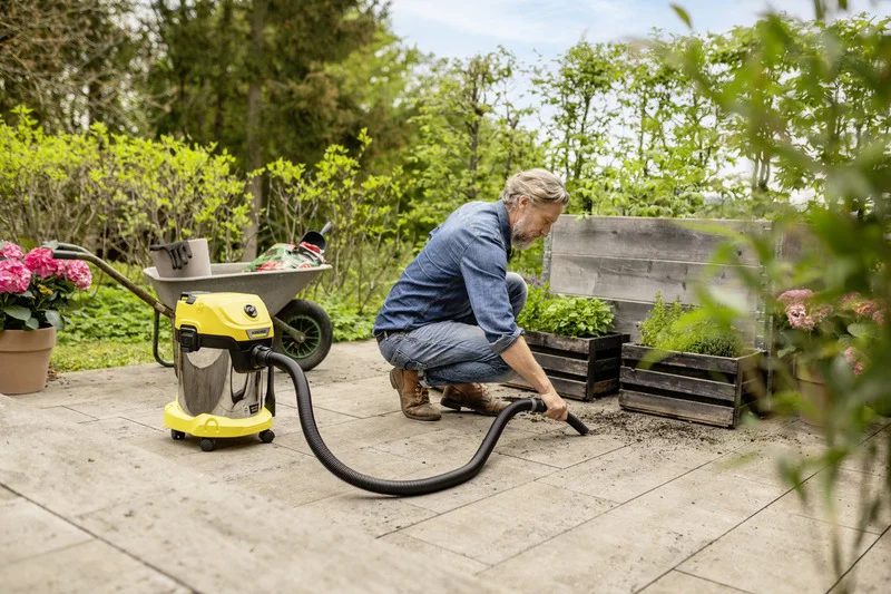 Person reinigt Terrasse mit Kärcher Nass-Trockensauger, neben Blumenkübel und Schubkarre, in einem Garten mit grünen Pflanzen.