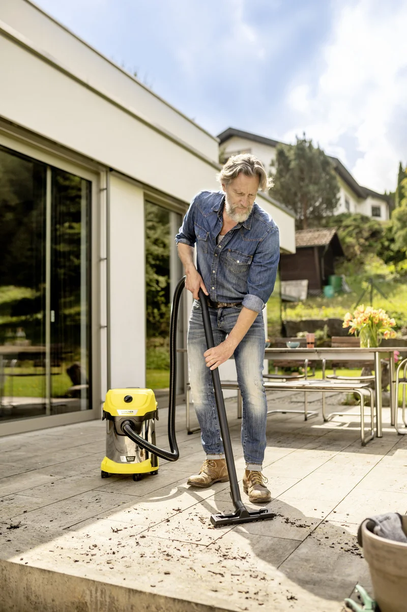 Person reinigt Terrasse mit Kärcher Nass-Trockensauger. Im Hintergrund ein Tisch mit Blumen und ein Haus. Outdoor-Szene bei Tageslicht.