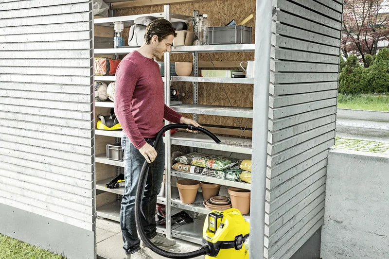 Mann in rotem Pullover hält Schlauch eines gelben Kärcher Nass-Trockensaugers in einem Gartenhaus mit Regalen voller Gartenzubehör.