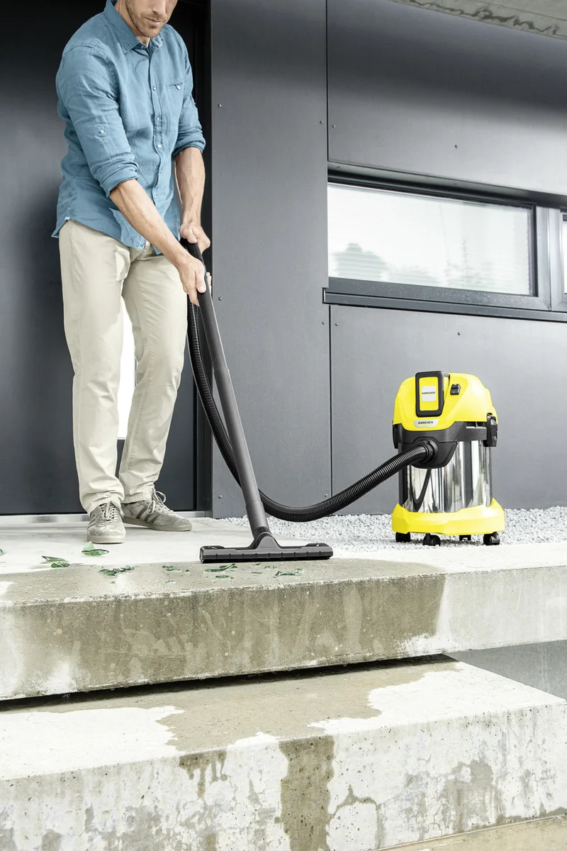 Mann reinigt Außentreppe mit einem Kärcher Nass-/Trockensauger. Betonstufen mit Wasserflecken und Blättern. Moderne Gebäudefassade im Hintergrund.