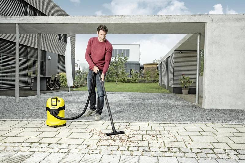 Person reinigt Terrasse mit Kärcher Nass-Trockensauger, umgeben von modernen Gebäuden und Garten im Freien.