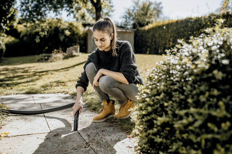 Person kniet im Garten und hält einen Kärcher Hochdruckreiniger, um eine gepflasterte Fläche zu reinigen. Im Hintergrund sind Bäume und ein Schuppen.