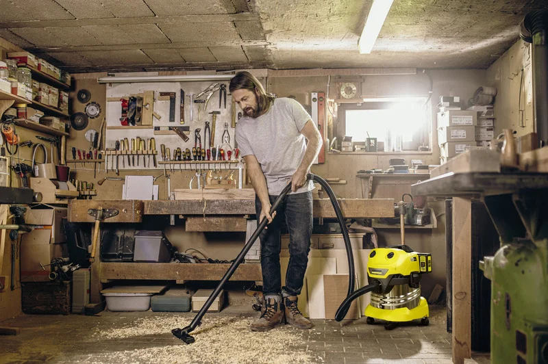 Mann reinigt Werkstattboden mit Kärcher Nass-/Trockensauger. Hintergrund zeigt Werkbank mit Werkzeugen und Regalen.