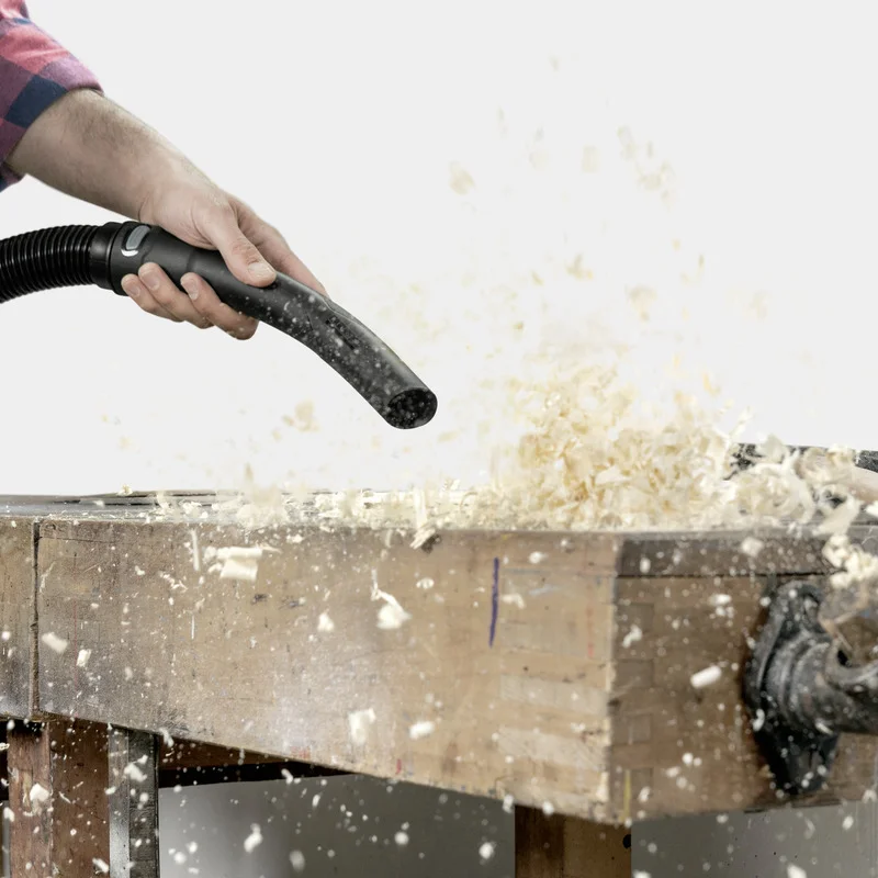 Hand hält Kärcher Staubsaugerrohr, saugt Holzspäne von Werkbank. Staub und Späne fliegen in der Luft.