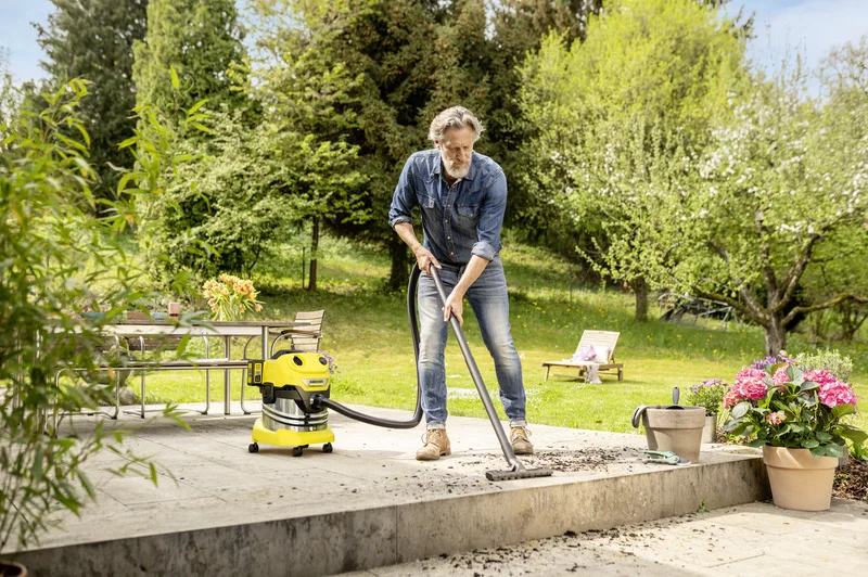 Person reinigt Terrasse mit Kärcher Nass-Trockensauger. Gartenmöbel und blühende Pflanzen im Hintergrund. Sonniger Tag im Freien.