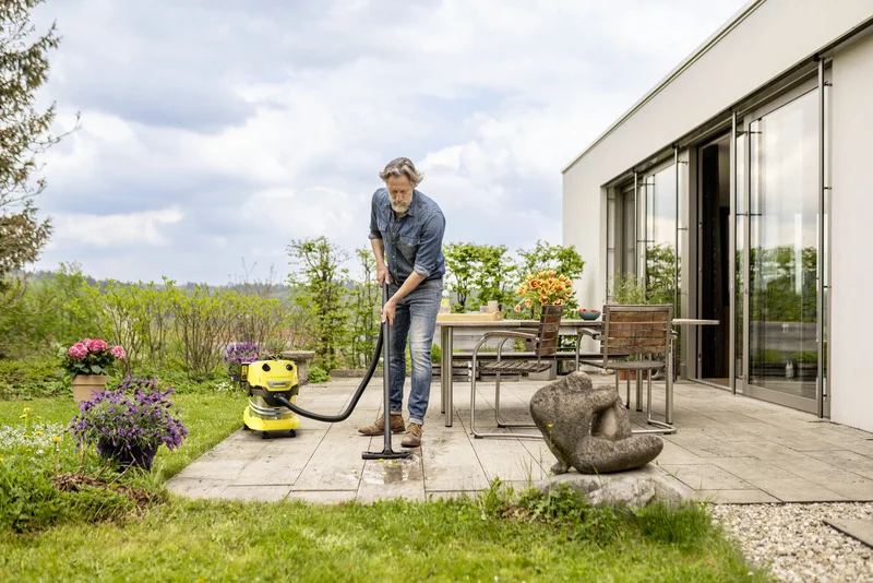 Mann reinigt Terrasse mit Kärcher Nass-Trockensauger. Im Hintergrund Gartenmöbel, Blumen und moderne Glasfassade.