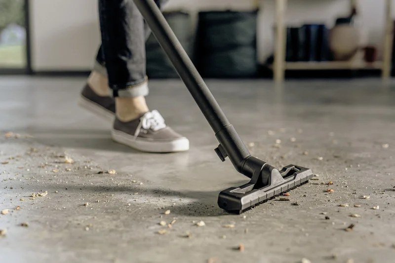 Person reinigt Boden mit Kärcher Staubsauger, trägt graue Schuhe. Staub und Schmutz auf Betonboden in Innenraum.