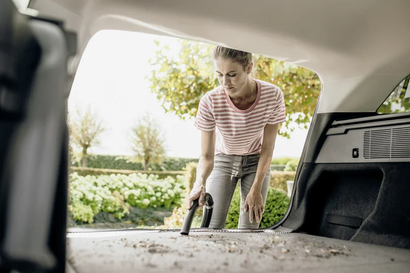 Person reinigt Kofferraum eines Autos mit einem Staubsauger, draußen im Garten mit Bäumen und Sträuchern im Hintergrund.