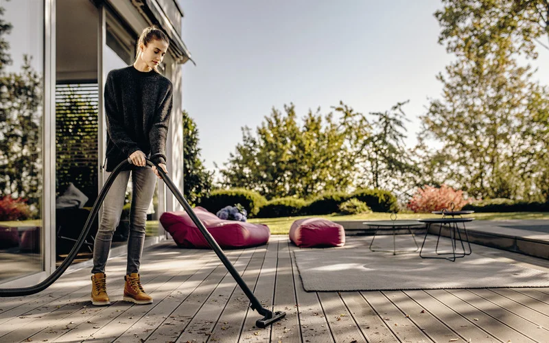 Person reinigt Terrasse mit Staubsauger, umgeben von Sitzsäcken und Tisch im Freien. Sonniger Tag, Bäume im Hintergrund.