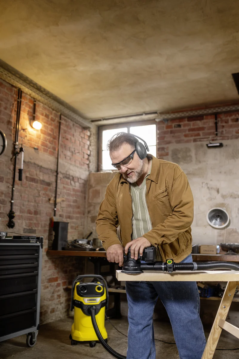 Mann in Werkstatt arbeitet mit Elektrowerkzeug an Holzplatte. Im Hintergrund steht ein gelber Kärcher Nass-Trockensauger.