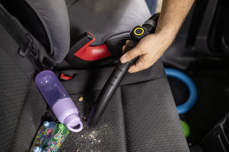 Hand hält Kärcher Staubsaugerdüse, reinigt Krümel auf Autositz neben lila Trinkflasche und buntem Spielzeug.