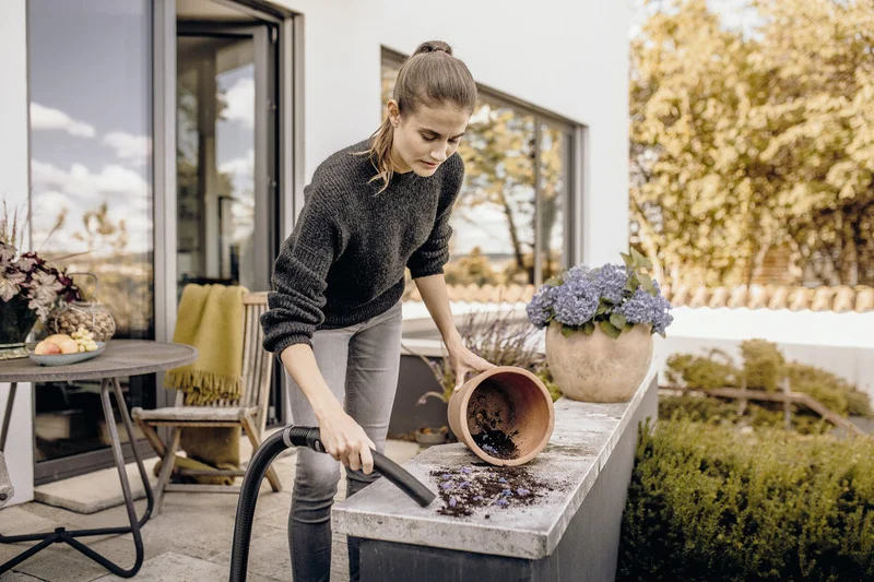 Person reinigt Terrasse mit Kärcher Staubsauger, Erde aus umgekipptem Blumentopf. Im Hintergrund Garten und moderne Hausfassade.