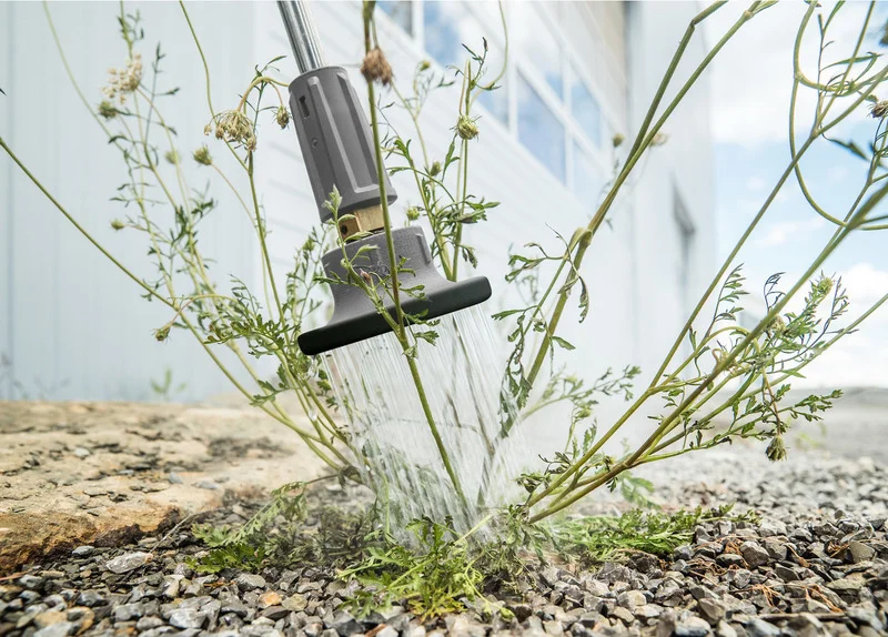 Kärcher Unkrautentferner sprüht Wasser auf Pflanzen zwischen Kiessteinen vor einem Gebäude.