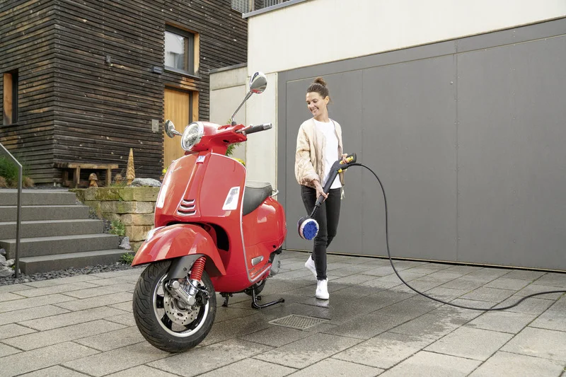 Rote Vespa auf gepflastertem Hof vor modernem Gebäude. Person hält Kärcher Hochdruckreiniger mit Bürstenaufsatz.