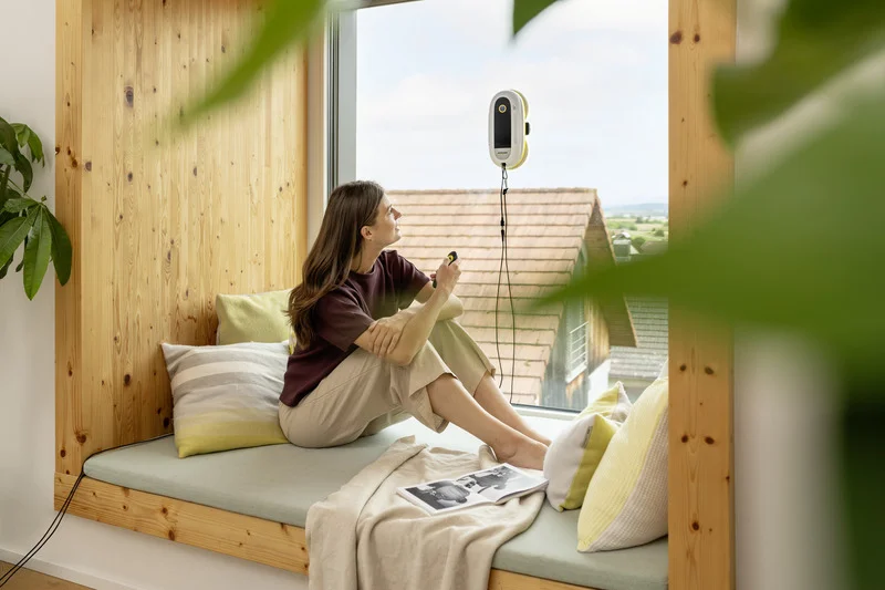 Frau sitzt auf Fensterbank, hält Smartphone, schaut nach draußen. Kärcher Fensterreiniger an der Scheibe. Innenraum mit Holzverkleidung.