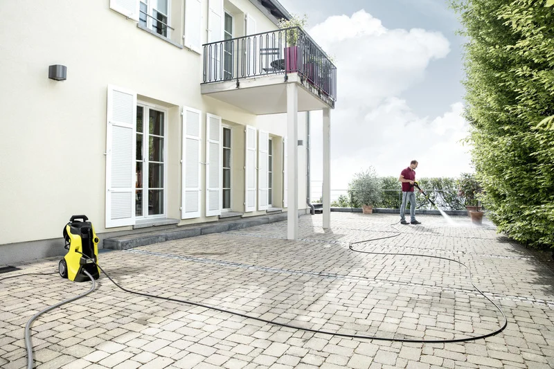 Mann reinigt Terrasse mit Kärcher Hochdruckreiniger. Gelbes Gerät steht auf gepflastertem Boden vor einem Haus mit weißen Fensterläden.