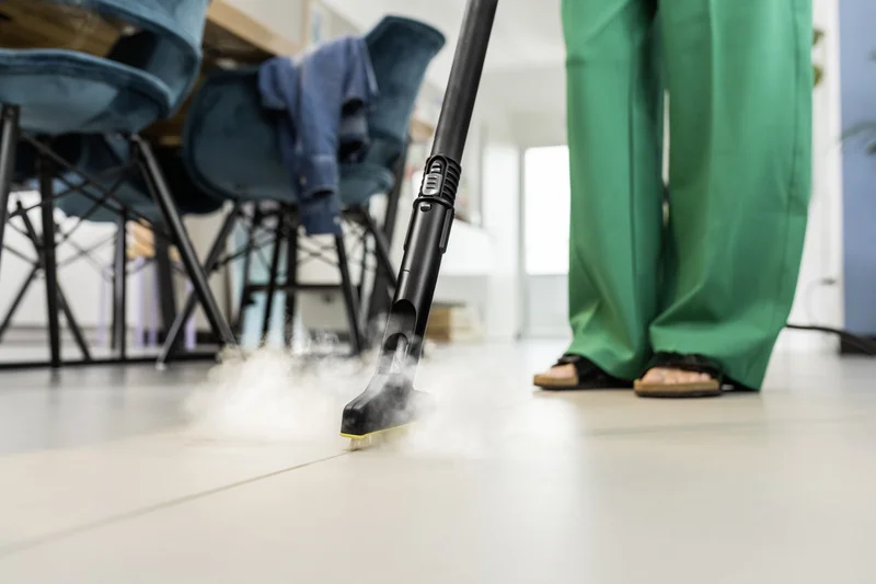 Person in grüner Hose bedient Kärcher Dampfreiniger auf Fliesenboden in modernem Raum mit blauen Stühlen.