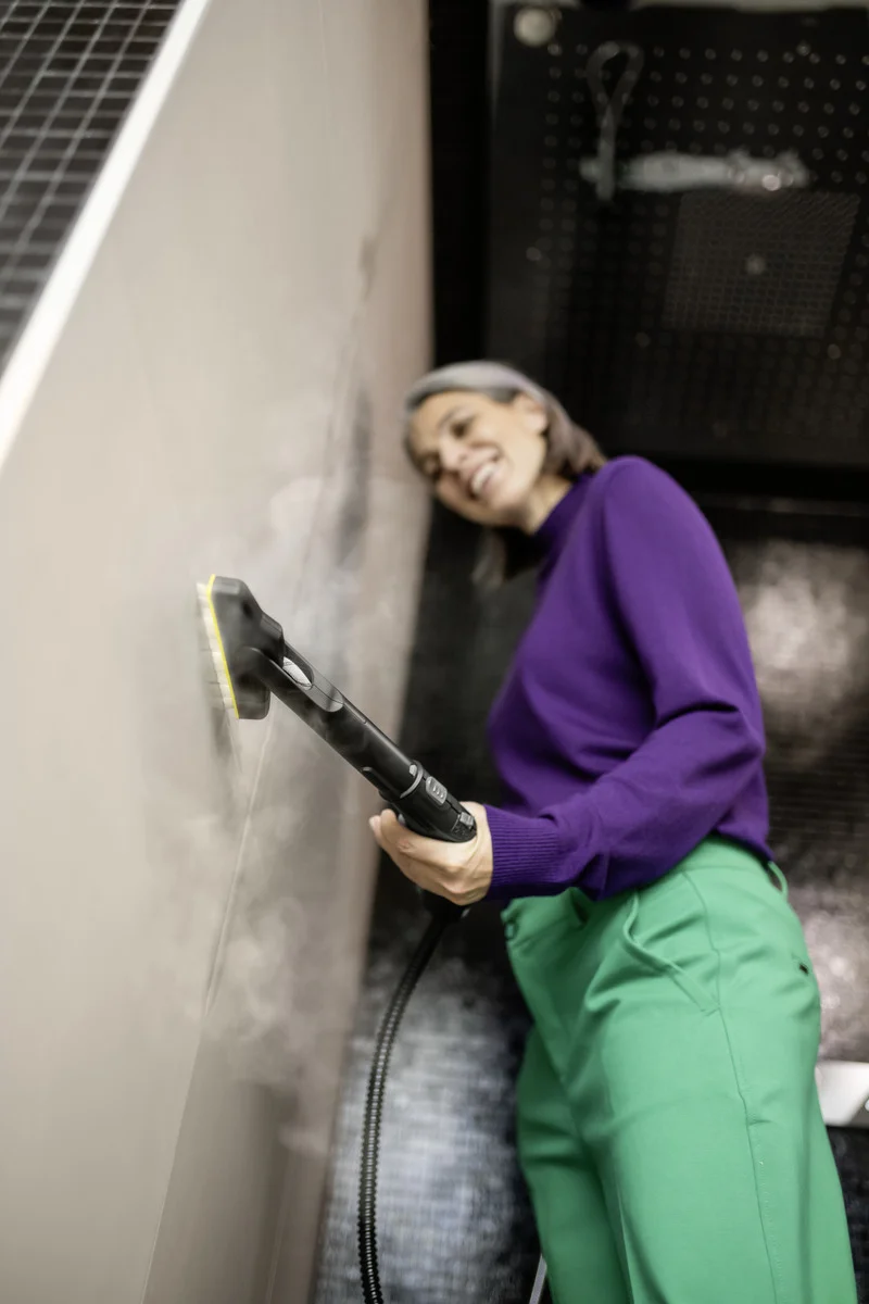 Person in lila Pullover und grüner Hose reinigt mit einem Kärcher Dampfreiniger eine Wand. Umgebung wirkt industriell.