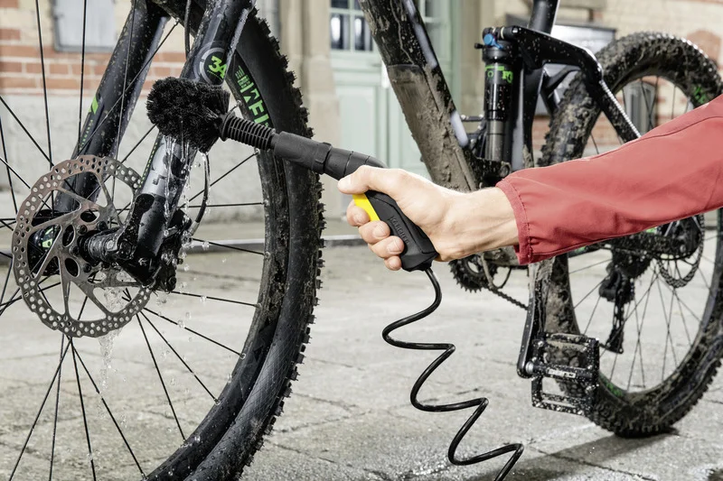 Hand reinigt Fahrradgabel mit Kärcher Reinigungsbürste. Fahrrad ist schlammig, Wasser tropft. Hintergrund: Gebäude mit Fenster.
