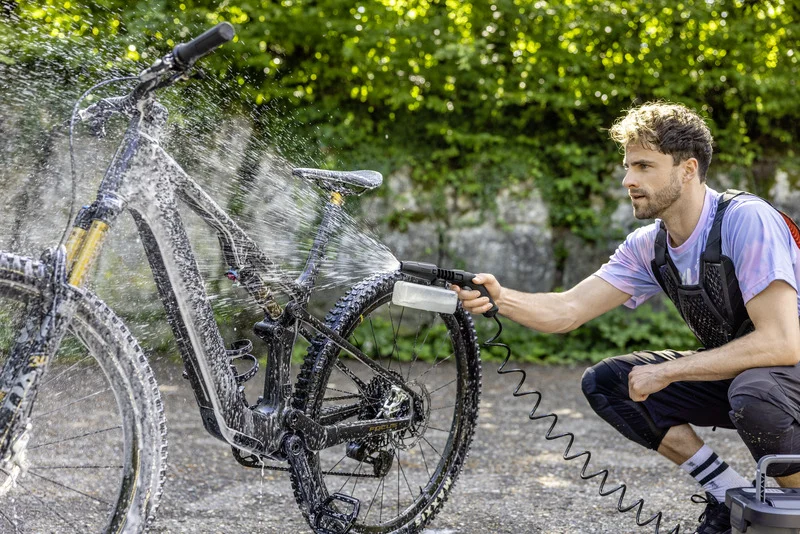 Person reinigt Fahrrad mit Kärcher Hochdruckreiniger im Freien, Wasser spritzt auf Rahmen und Reifen, grüne Hecke im Hintergrund.