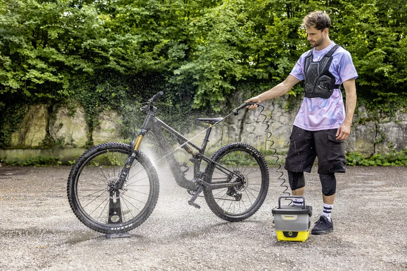 Person reinigt Fahrrad mit Kärcher Hochdruckreiniger im Freien, umgeben von Bäumen und Steinmauer.