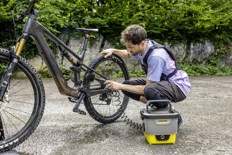 Person reinigt Fahrrad mit Kärcher Mobile Outdoor Cleaner auf einem Parkplatz, umgeben von Bäumen und einer Steinmauer.