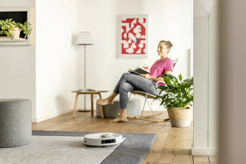 Saugroboter auf Teppichboden in modernem Wohnzimmer. Person sitzt lesend im Schaukelstuhl, umgeben von Pflanzen und Kunst an der Wand.