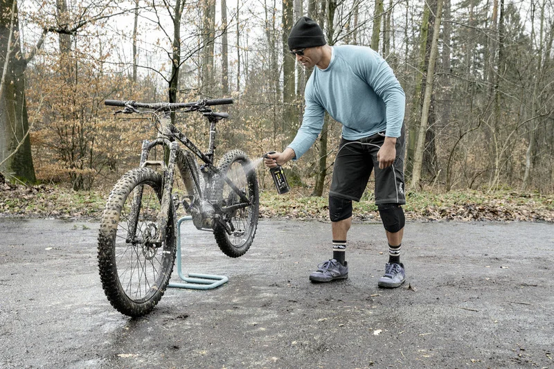 Mann reinigt schmutziges Mountainbike im Wald mit einem Reinigungsmittel aus einer Sprühflasche. Fahrrad steht auf einem Ständer.