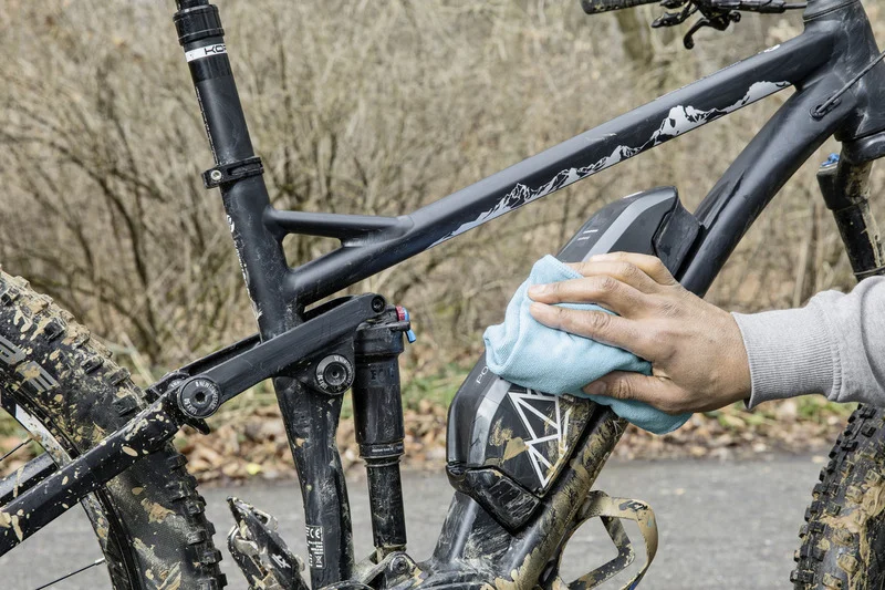 Hand reinigt ein schmutziges Mountainbike mit einem blauen Tuch im Freien. Hintergrund zeigt kahle Bäume.
