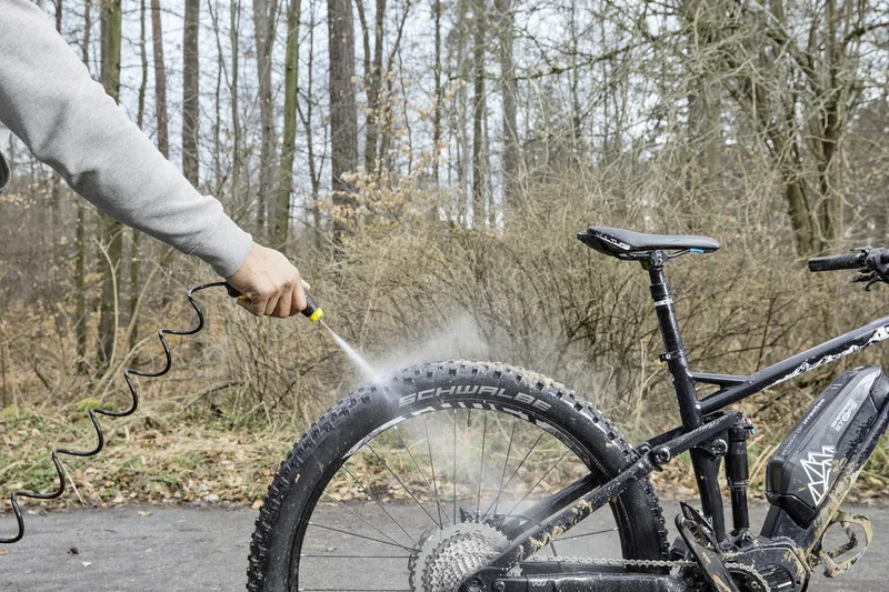 Person reinigt Fahrradreifen mit Kärcher Hochdruckreiniger im Wald. Fahrrad ist schlammig, Umgebung mit Bäumen und Sträuchern.