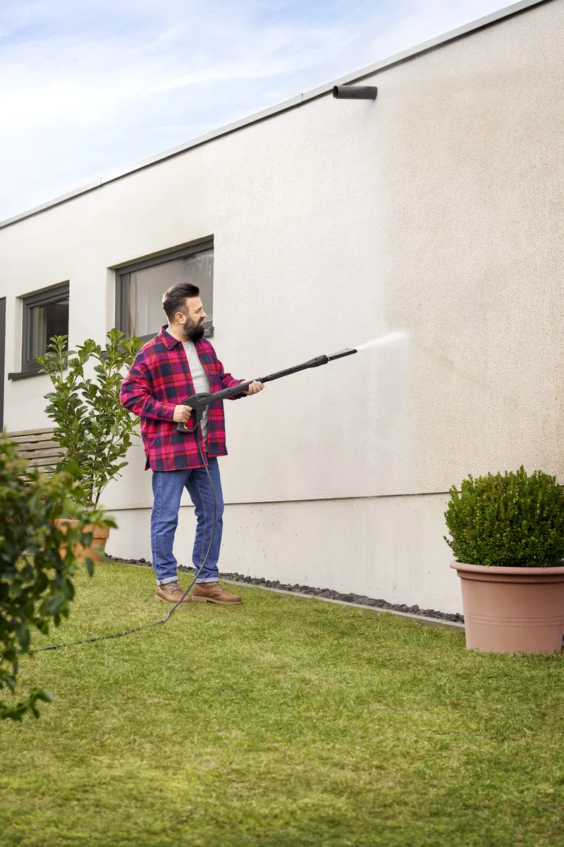 Person reinigt Hauswand mit Kärcher Hochdruckreiniger im Garten. Rote Karo-Jacke, Jeans, Pflanzen im Hintergrund.